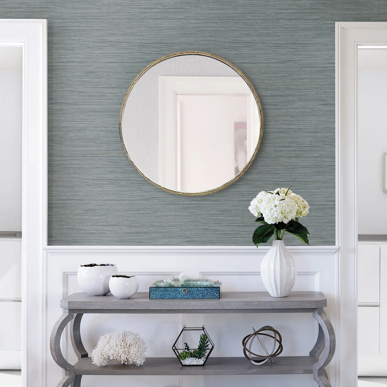 Textured blue wallpaper above white paneling, with a round mirror and console table